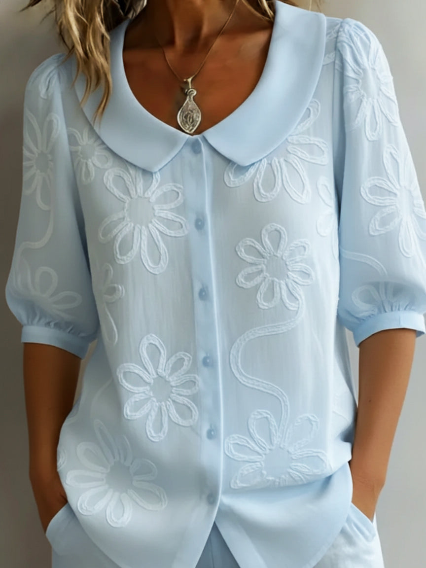 Blusa ligera azul claro con flores bordadas, cuello amplio y botones, manga tres cuartos.
