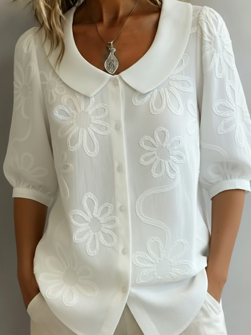 Blusa blanca ligera con flores bordadas, cuello amplio y botones, manga tres cuartos.